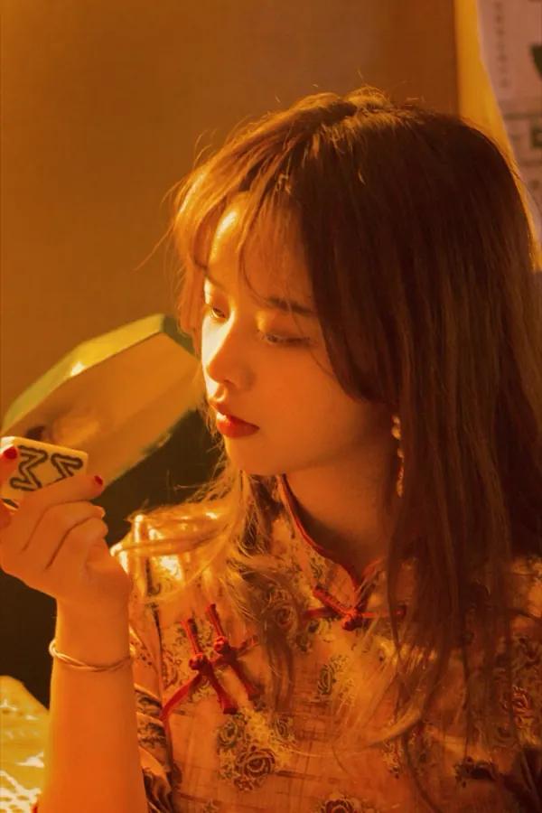A portrait in warm amber light: a qipao, and a mahjong tile held up close.