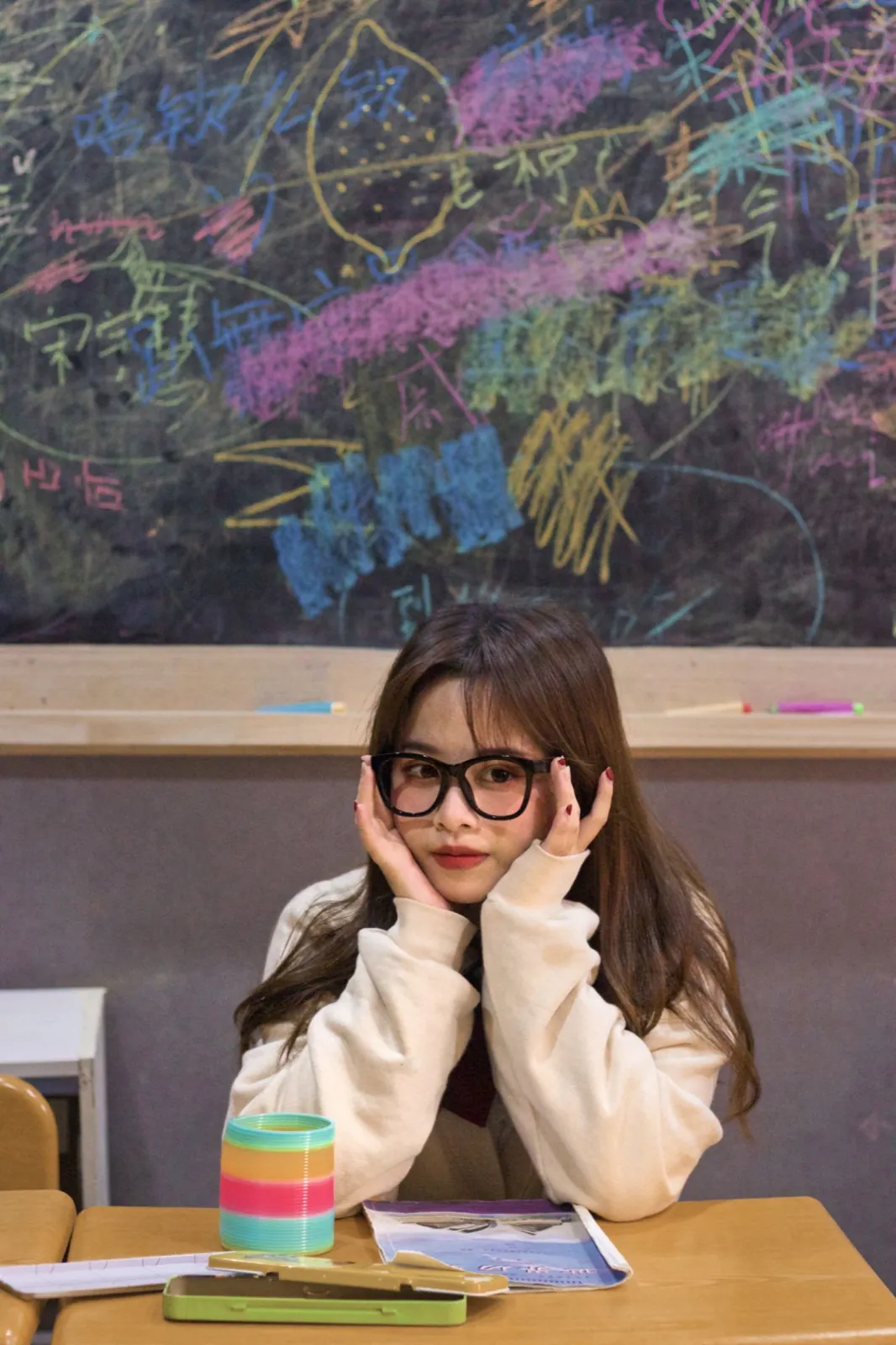 At a classroom desk in thick glasses, before a chalkboard full of coloured scribbles.
