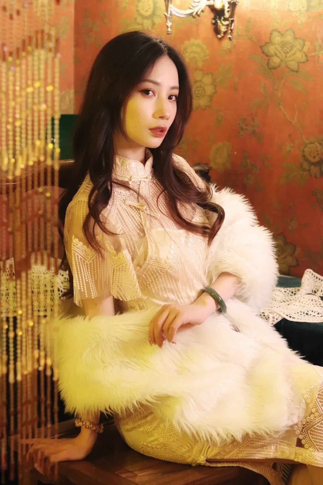 A cream lace qipao and a white fur stole, against floral wallpaper.